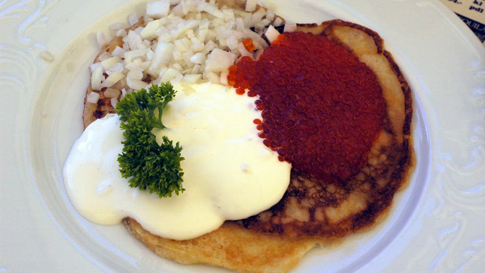 Blini med rød rogn, hakket løg, creme fraiche og persille på en hvid tallerken