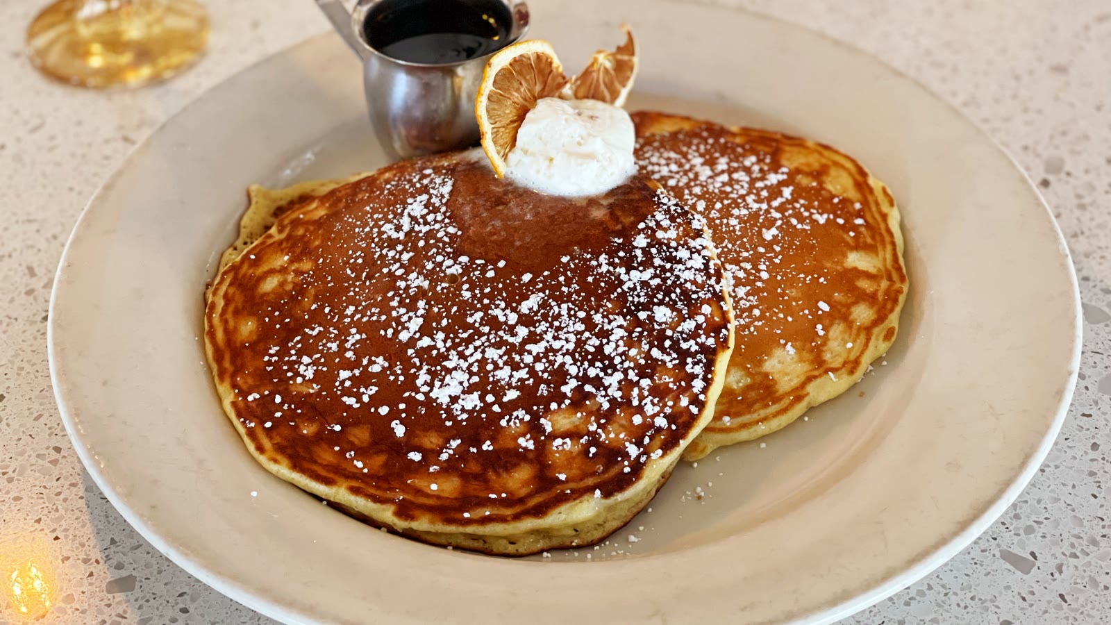 To pandekager med flormelis, en klat creme og en tørret citrusskive, med en lille kande sirup ved siden af